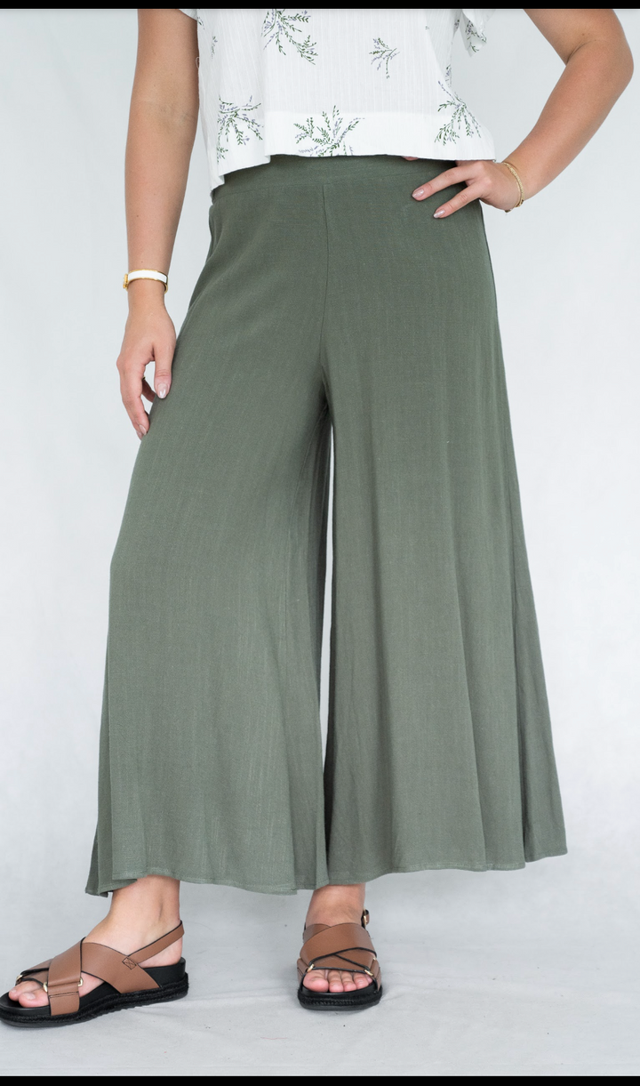 A model wearing high-waisted, wide-leg olive green pants, styled with a white sleeveless top and brown strappy sandals against a plain light gray background.