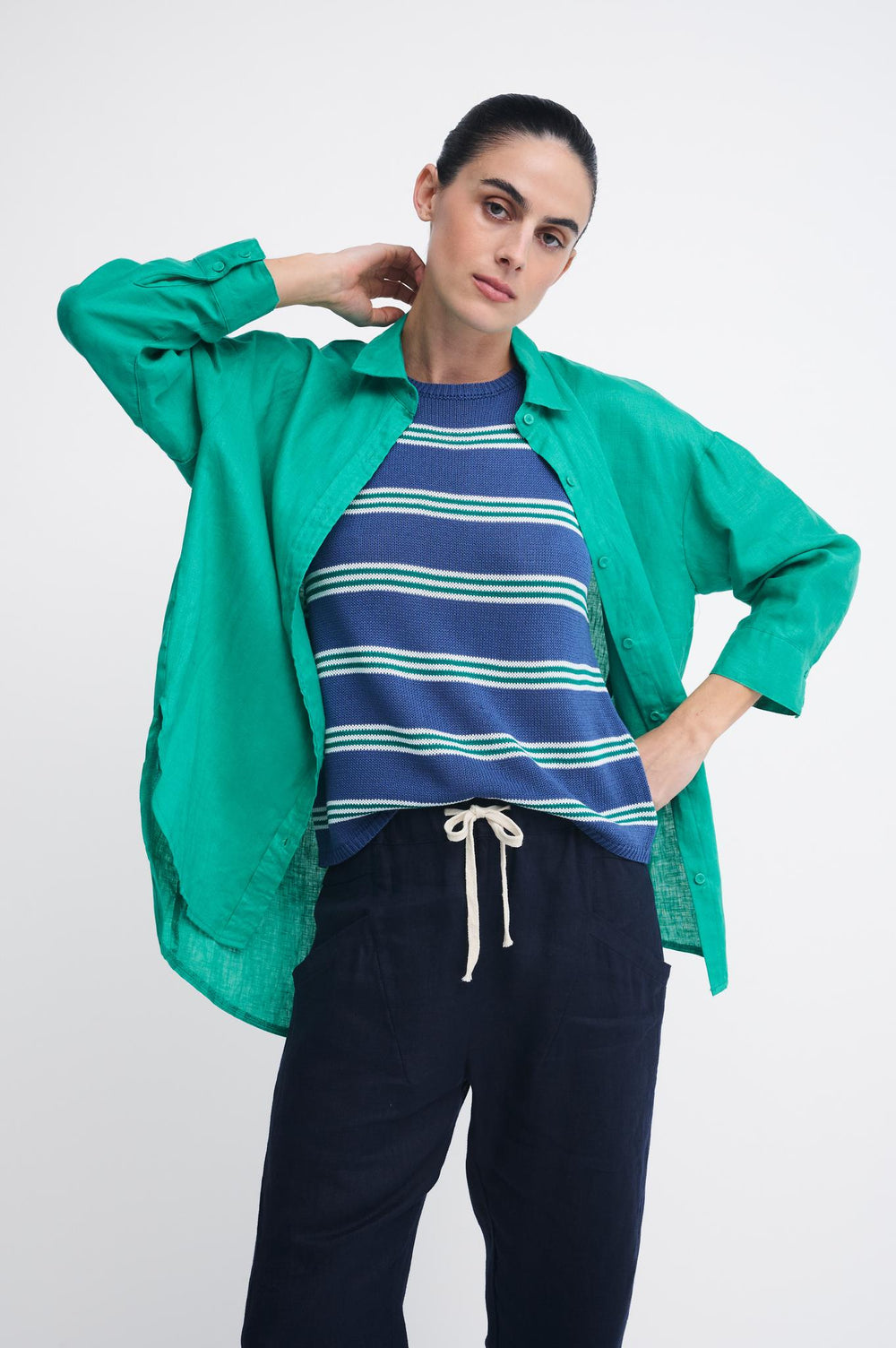 A model posing in a bright green linen button-up shirt layered over a navy blue and green striped t-shirt, paired with dark navy drawstring pants, against a light gray studio background.