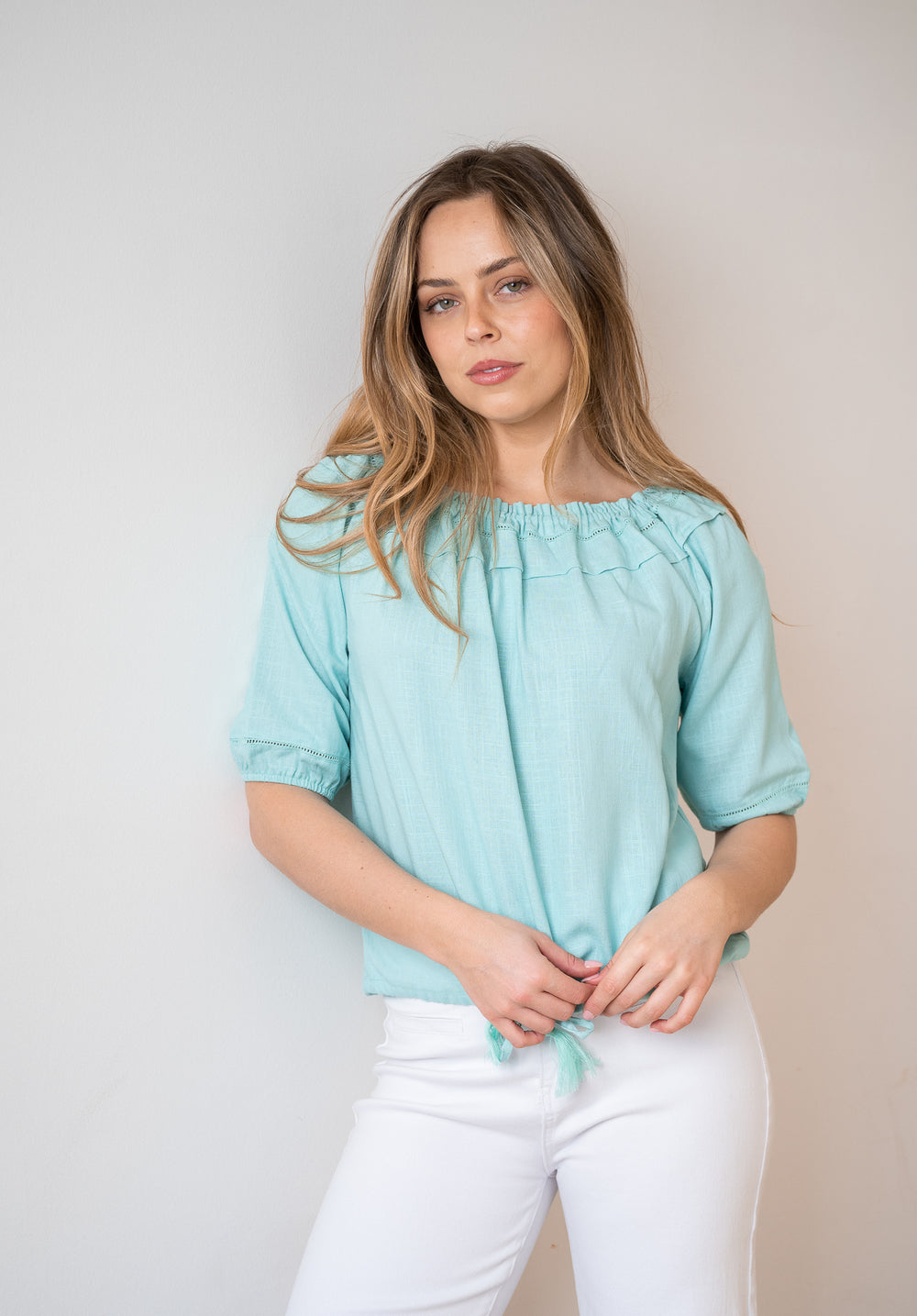 A woman wearing a light mint green short-sleeve blouse with a gathered neckline and white pants, standing against a plain light gray wall.