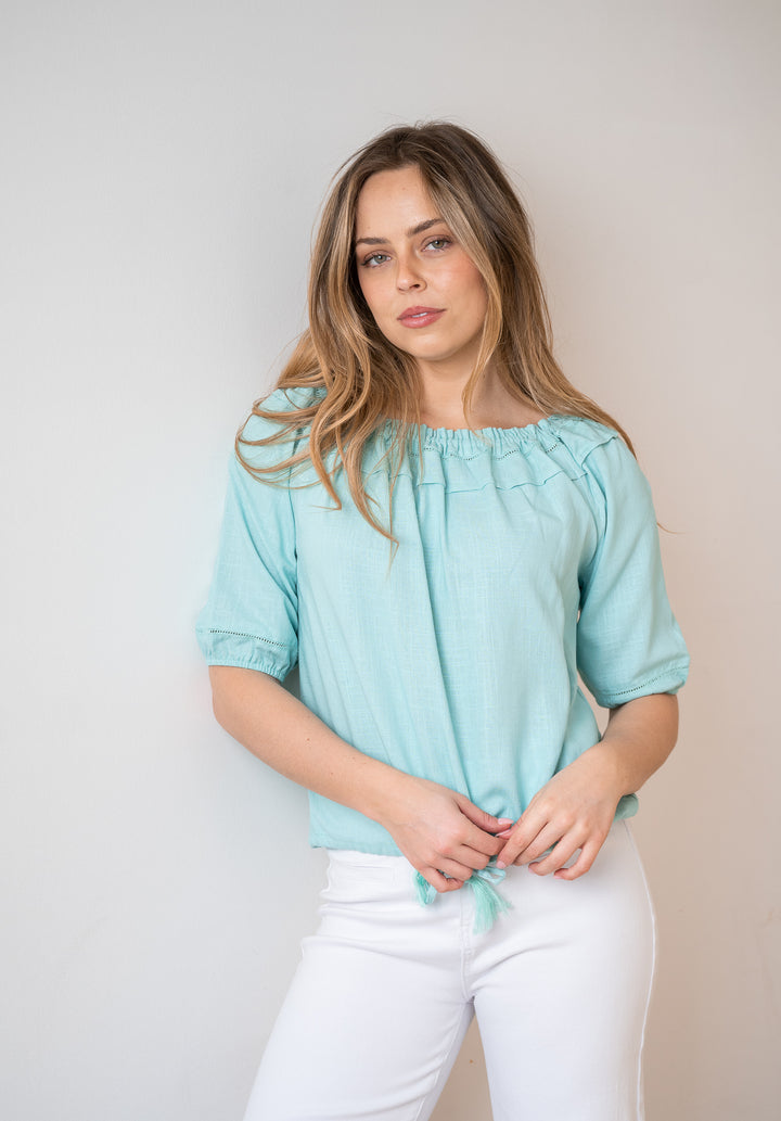 A woman wearing a light mint green short-sleeve blouse with a gathered neckline and white pants, standing against a plain light gray wall.