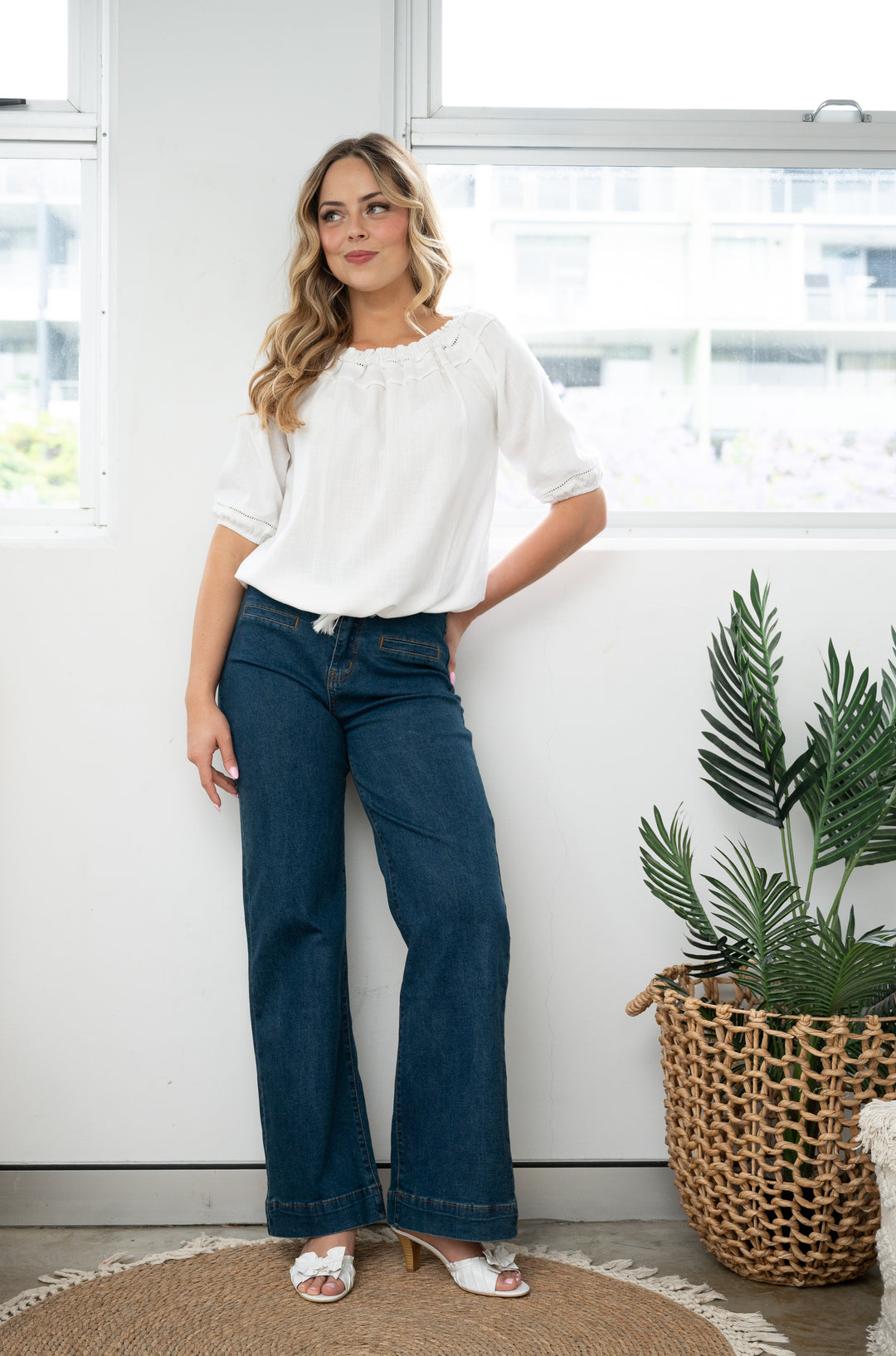 Woman wearing a white blouse and blue jeans standing in a bright room with a plant.