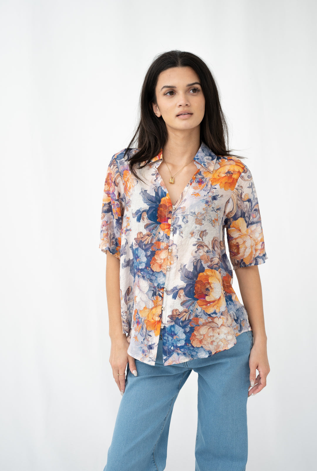 Woman wearing a short-sleeve floral blouse in blue, amber, and cream tones, styled with light blue jeans against a white background