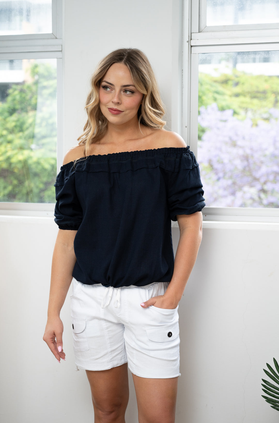Woman wearing a NAVY off-shoulder top and white shorts standing indoors with a window in the background.