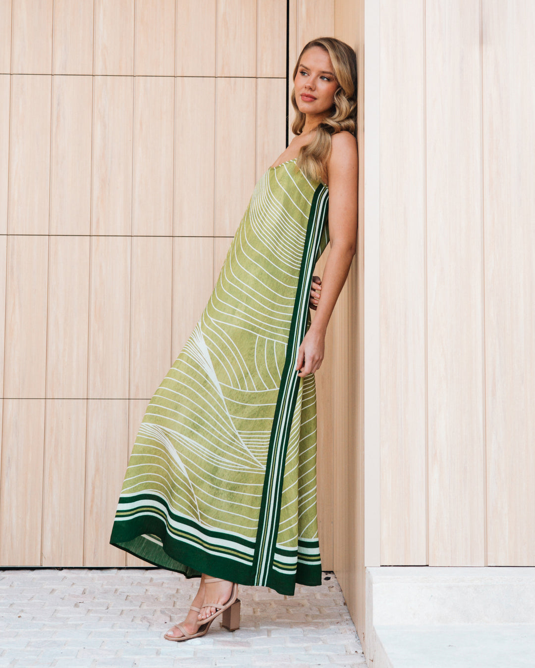 A woman models a sleeveless maxi dress with a green and white wavy line pattern, leaning against a light wood-paneled wall.