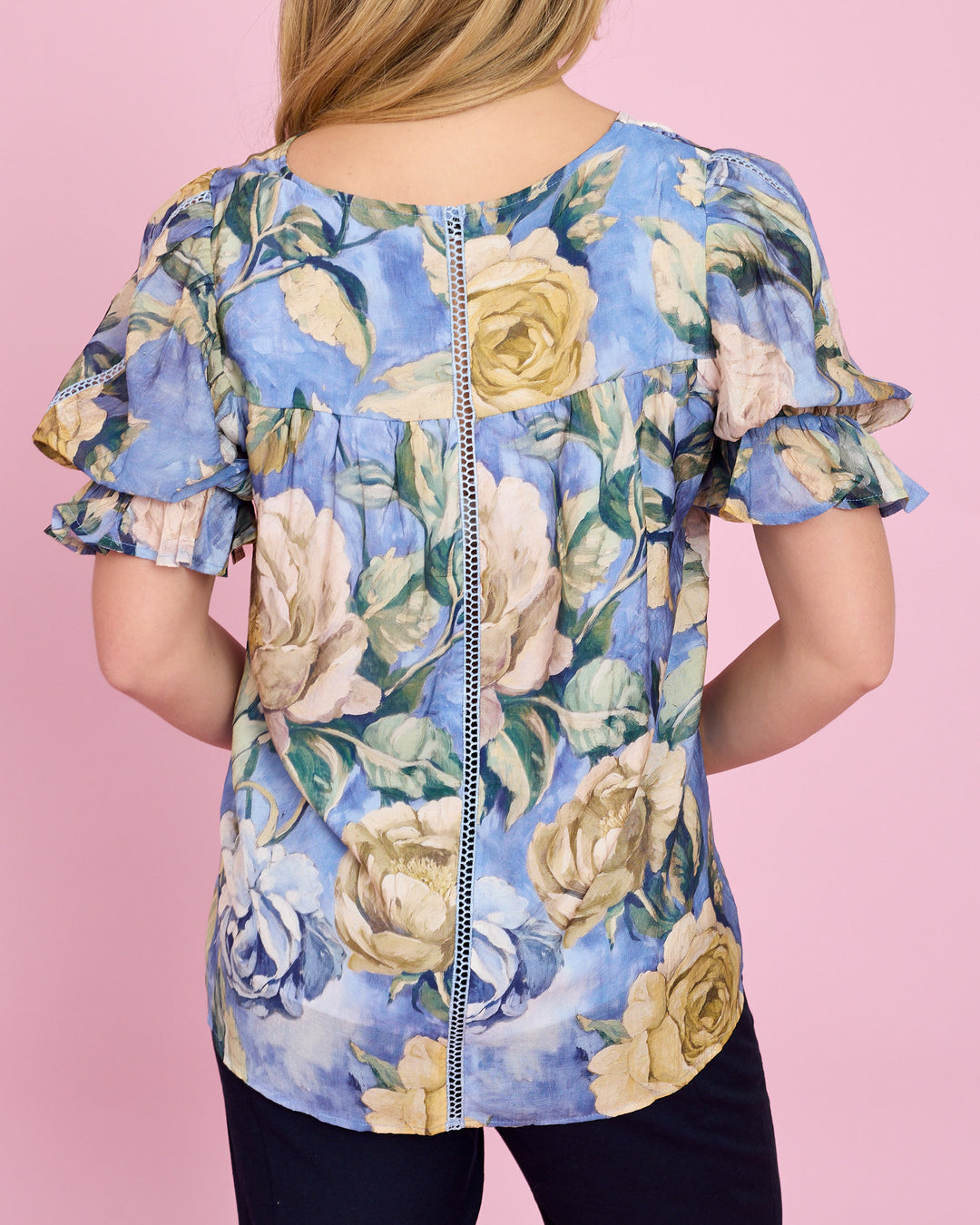A woman shown from the back wearing a short-sleeve ruffled blouse with a blue watercolor floral print featuring cream and yellow roses, set against a solid pink background.