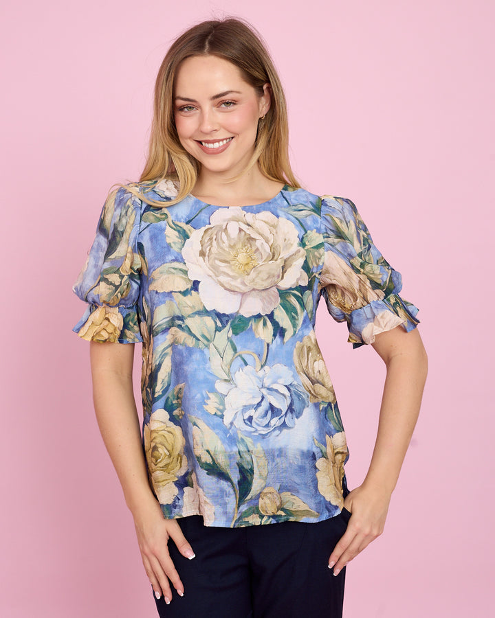 A woman modeling a short-sleeve blouse with a blue floral print, featuring large cream and yellow roses with green leaves, set against a solid pink background.