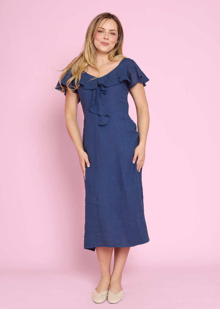 A woman modeling a blue midi dress with ruffled short sleeves and a tie detail at the neckline, standing against a solid pink studio background.