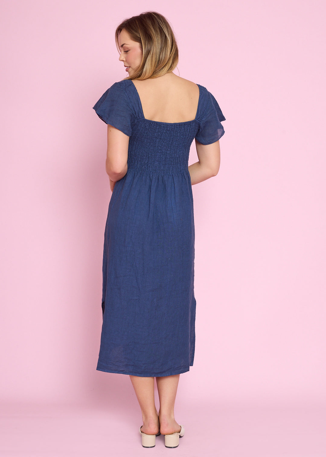 A woman wearing a smocked midi dress in a dusty blue shade, shown from the back against a solid pink studio background.