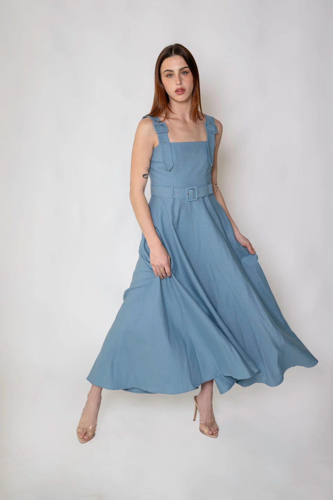 A woman modeling a dusty blue sleeveless maxi dress with shoulder straps, a matching belt, and side pockets, standing against a light gray studio background.