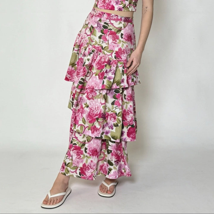A model wearing a pink and green floral tiered maxi skirt with a matching crop top, styled with white flip-flop sandals against a plain light gray background.