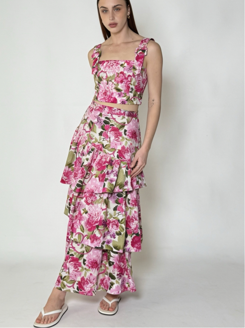 A woman modeling a two-piece floral set consisting of a sleeveless crop top and a matching tiered maxi skirt. The set features a vibrant pink and green floral print on a white background, photographed against a plain light gray studio backdrop.