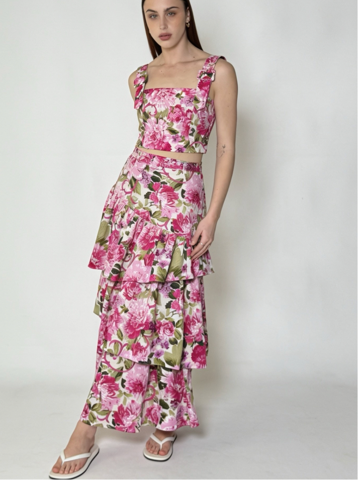 A woman modeling a two-piece floral set consisting of a sleeveless crop top and a matching tiered maxi skirt. The set features a vibrant pink and green floral print on a white background, photographed against a plain light gray studio backdrop.