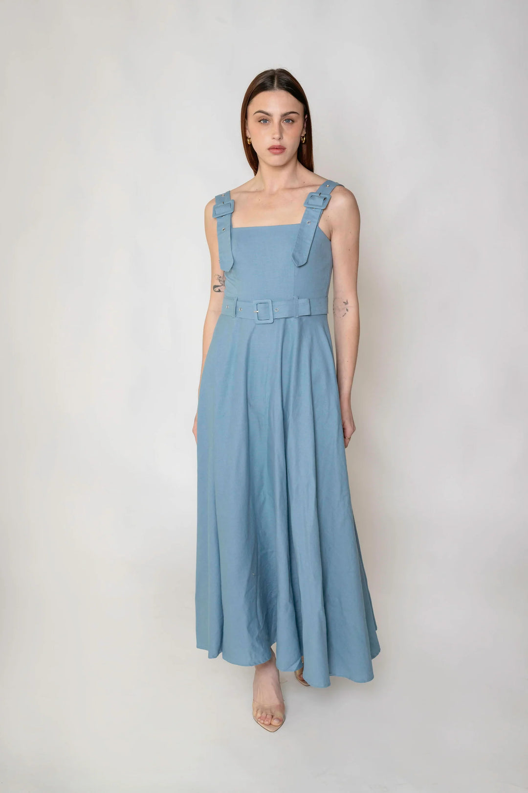 A woman modeling a light blue linen maxi dress with ruffled shoulder straps and a matching belt, standing against a neutral light gray background.