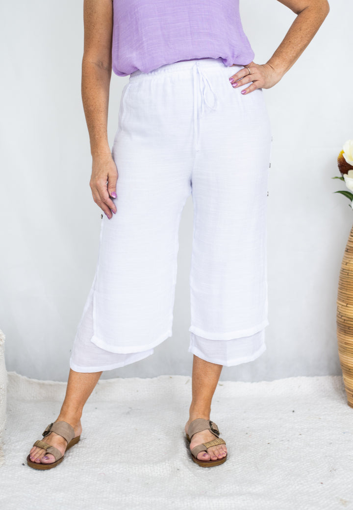 A person wearing loose white cropped pants with a drawstring waist, paired with a lavender top and brown sandals, standing against a light gray wall.