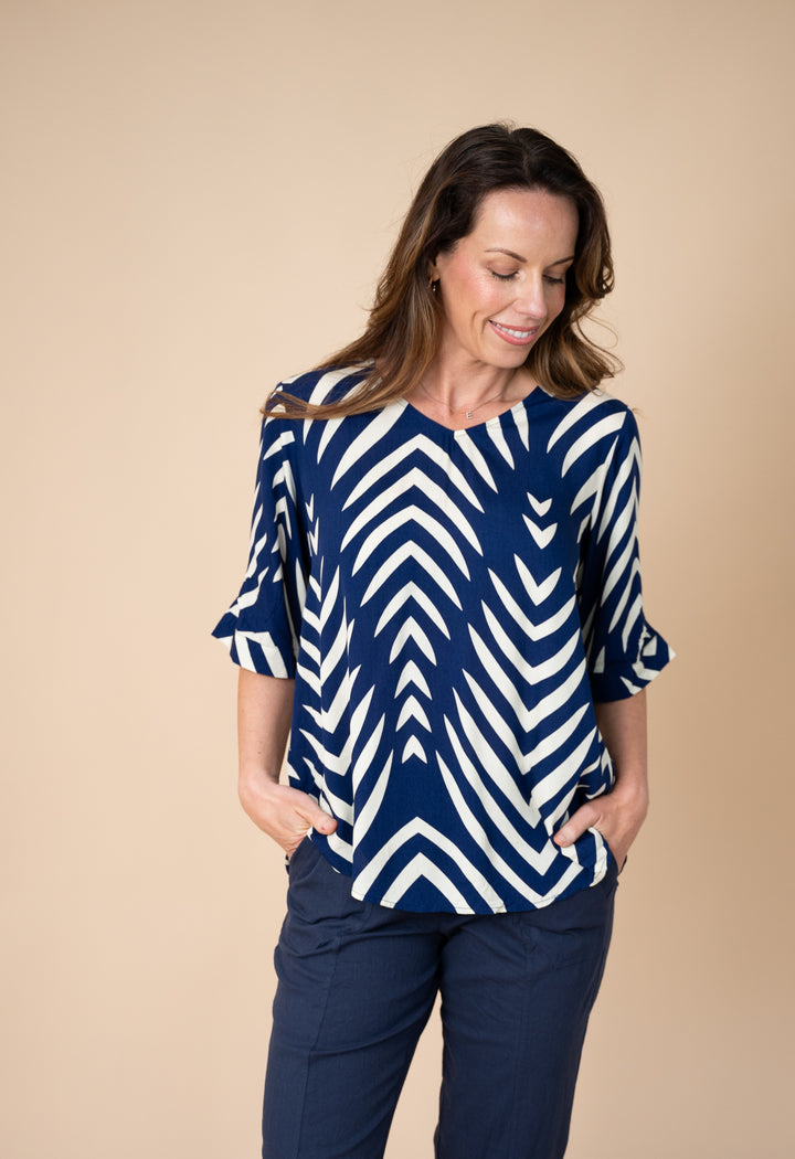 A woman wearing a navy blue and white chevron-patterned top with three-quarter ruffled sleeves, paired with navy pants, standing against a solid beige studio background.