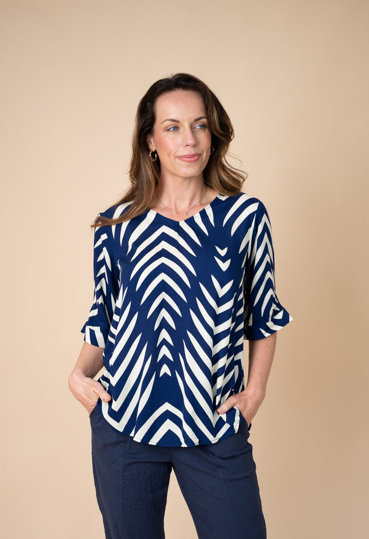 A woman modeling a navy blue and white geometric patterned top with three-quarter sleeves, paired with navy pants, against a solid beige studio background.