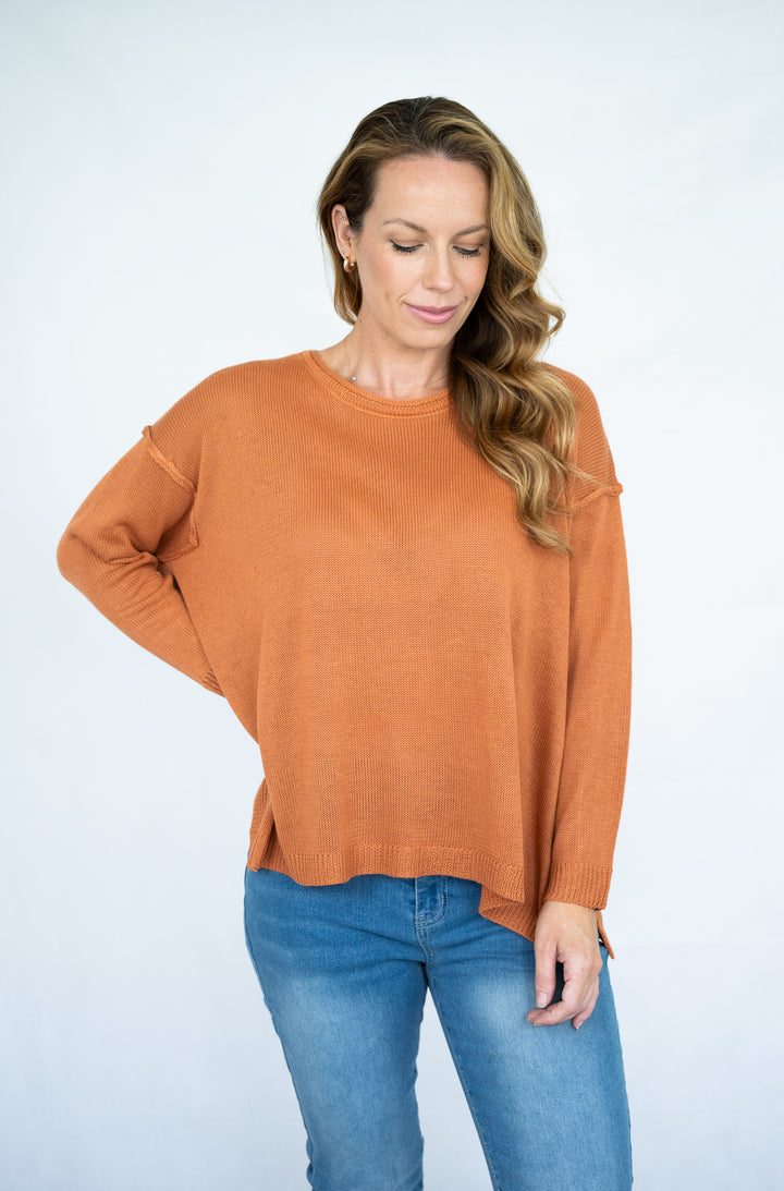 A woman wearing a rust-orange long-sleeve knit sweater and blue jeans, standing against a light gray studio background.