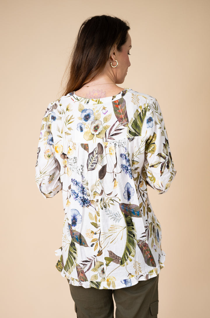 A woman viewed from the back wearing a white long-sleeve blouse with a colorful botanical print of blue, green, and yellow leaves, paired with olive green pants, against a solid beige background.