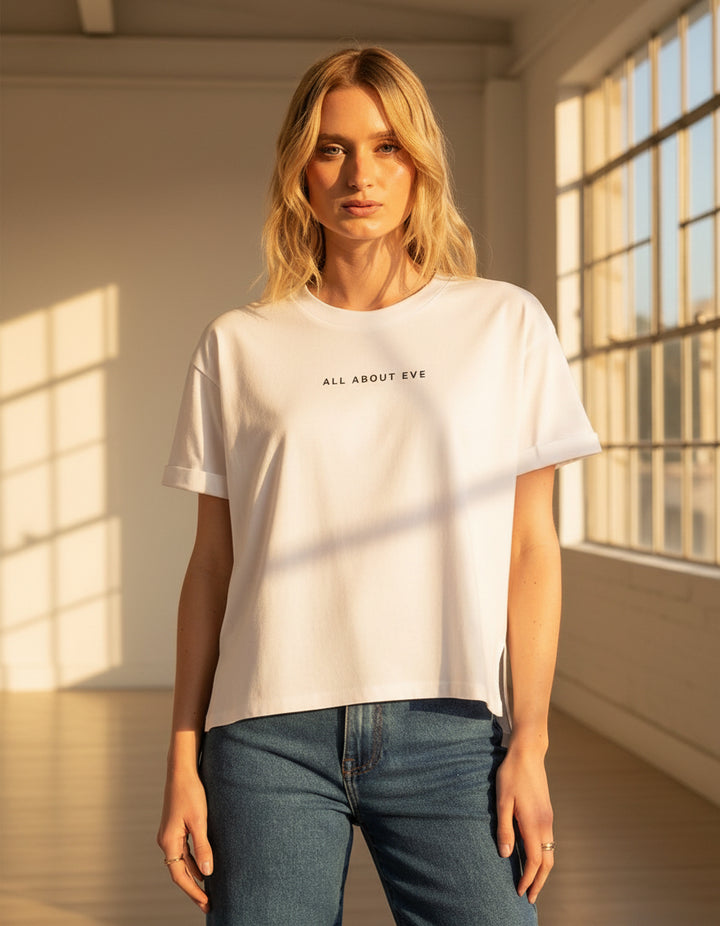 Woman wearing a white 'All About Eve' t-shirt and blue jeans on a light gray background
