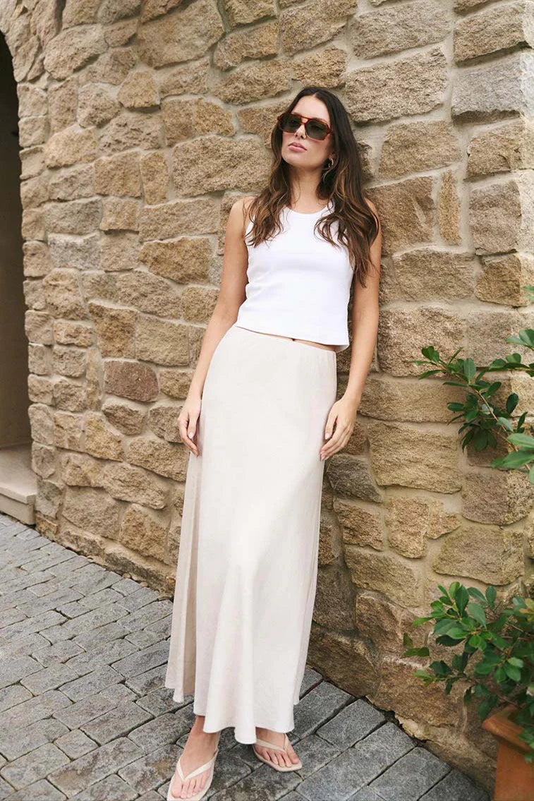 Woman in a white sleeveless top and long white skirt standing against a stone wall.
