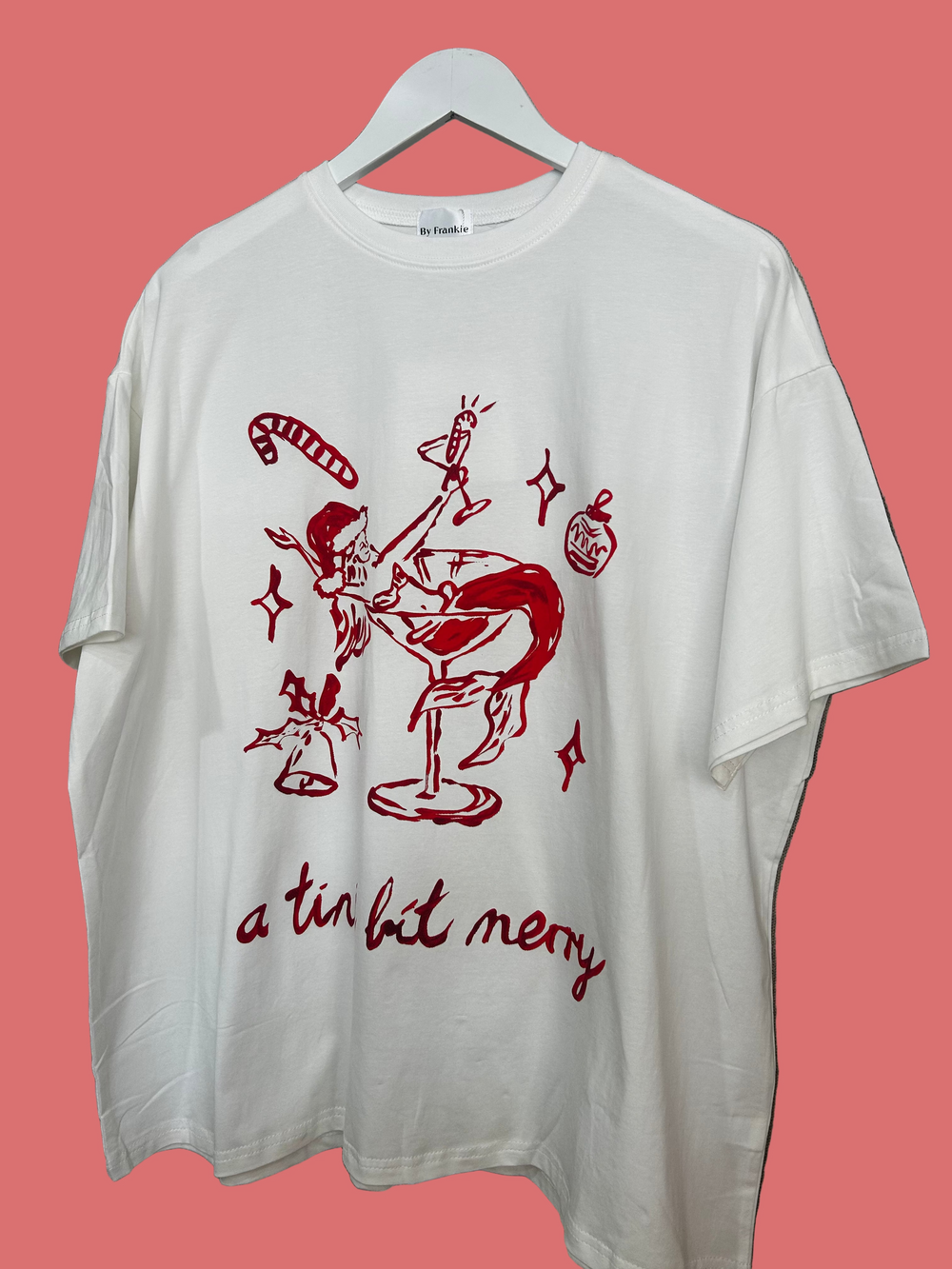 White t-shirt with red graphic design and text on a hanger against a neutral background White oversized Christmas graphic tee with red martini illustration and ‘a tini bit merry’ text by By Frankie