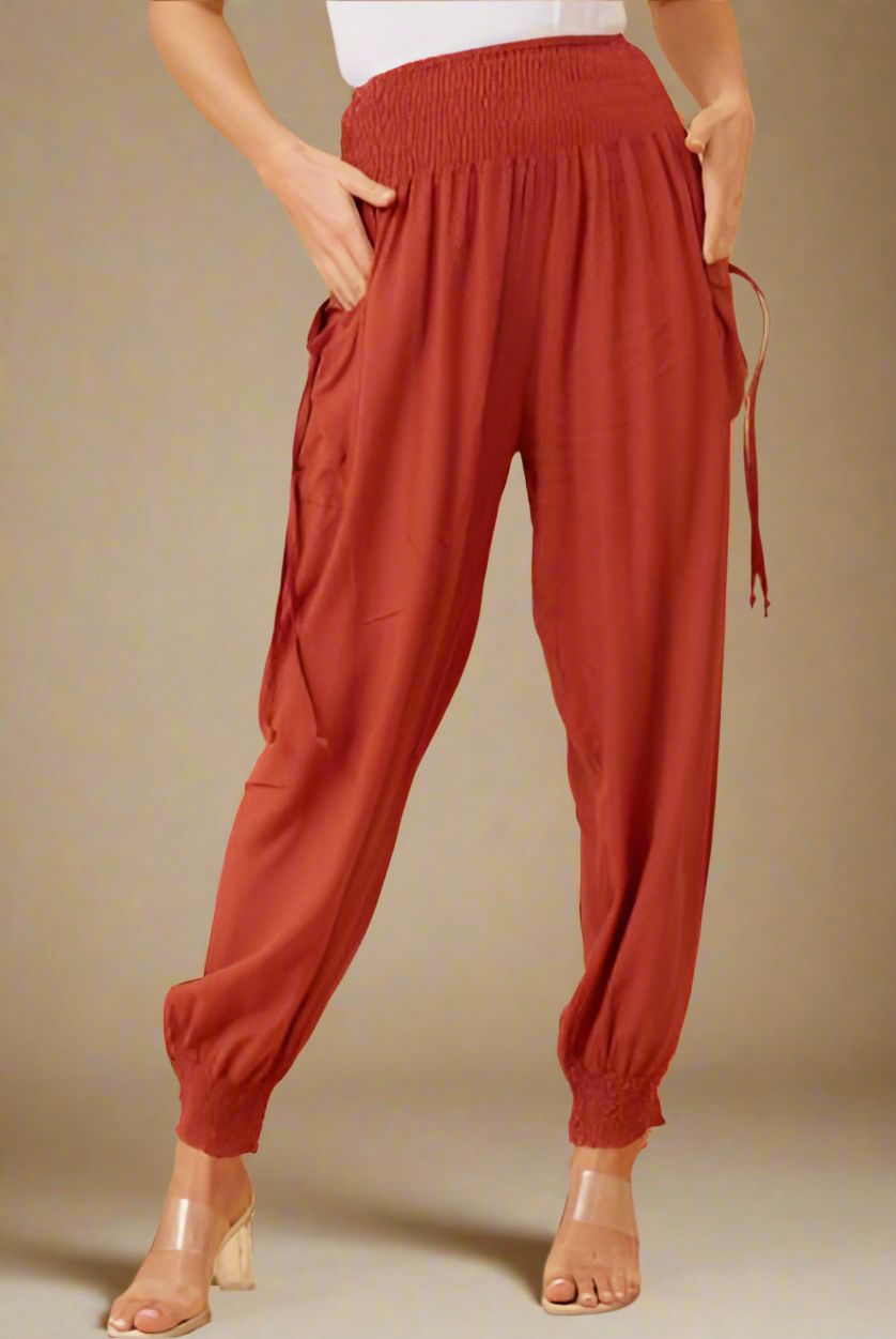 A person modeling loose, high-waisted harem-style pants in a rust red color, paired with a white top and clear high-heeled sandals, against a plain beige studio background.