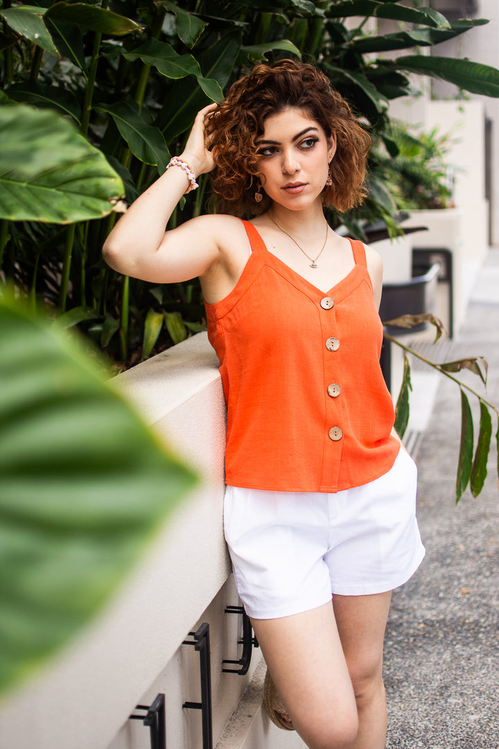 Woman wearing an orange sleeveless top and white shorts standing outdoors with greenery in the background
