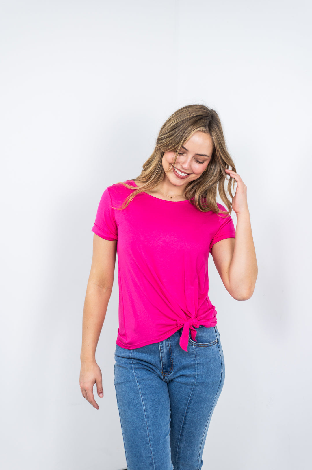 Woman wearing a bright pink t-shirt with a knot at the waist and blue jeans on a white background