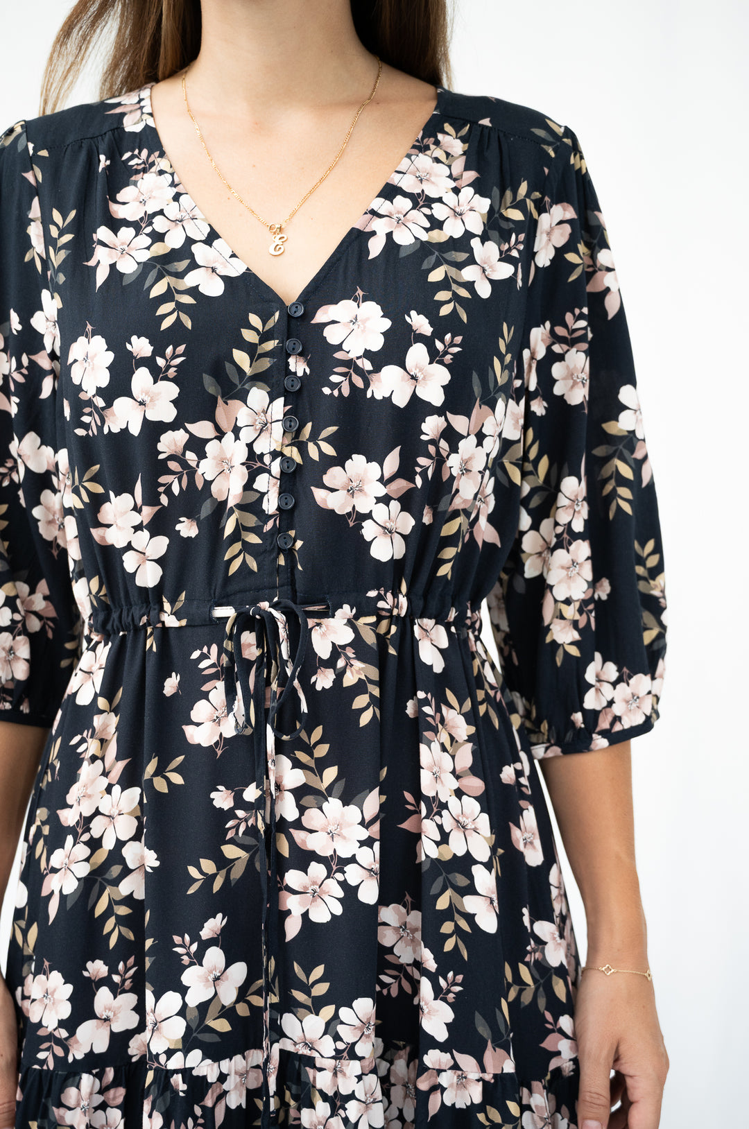 A woman wearing a black V-neck dress with a white and pink floral print, featuring elbow-length sleeves and a drawstring waist, shown against a plain white background.