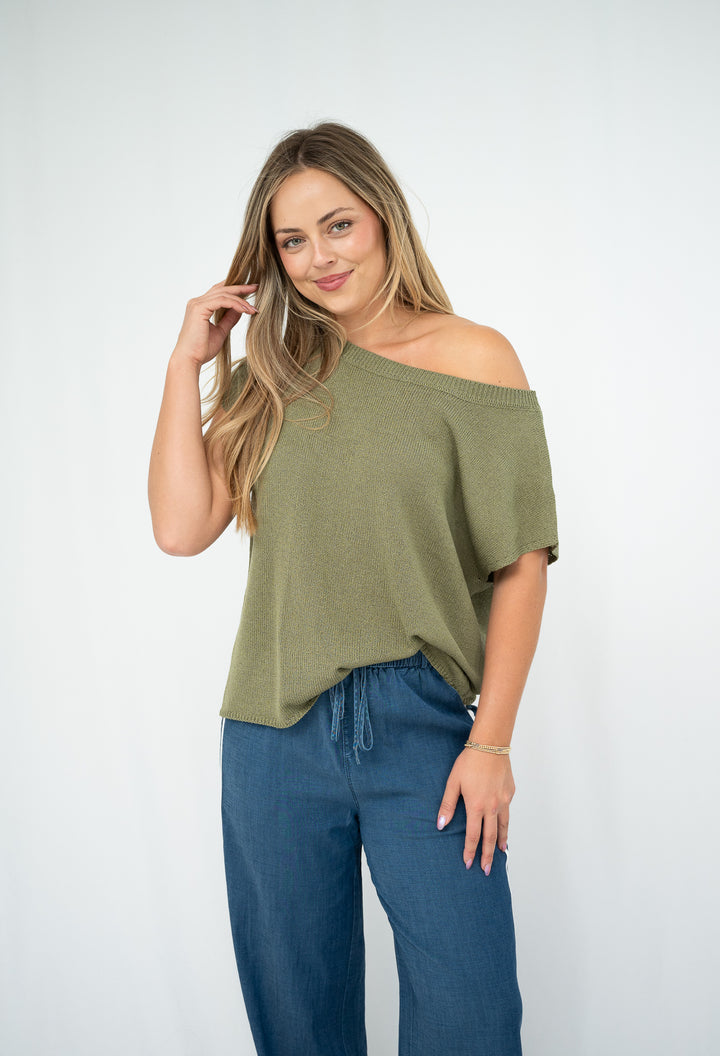 Woman wearing an olive green off-shoulder top and blue pants on a white background