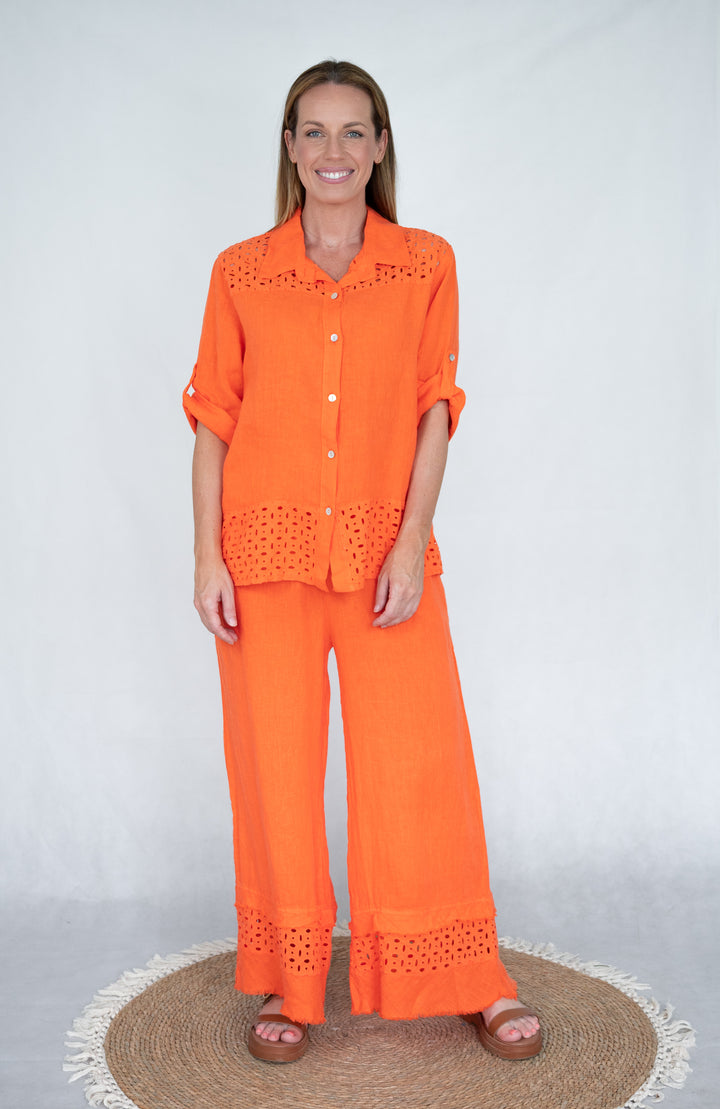 A woman standing, wearing an orange linen lattice shirt with button-up front and cut-out details on the shoulder and hem, paired with matching pants.