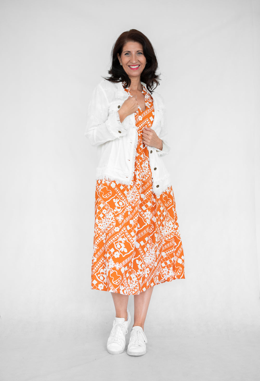 A person wearing a linen fringe jacket with a white and orange geometric patterned dress, paired with white shoes.